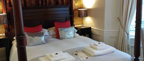 Un lit à baldaquin avec une structure en bois foncé, du linge de lit blanc et des coussins rouges et argentés. Il y a des serviettes sur le lit et une lampe sur la table de chevet.