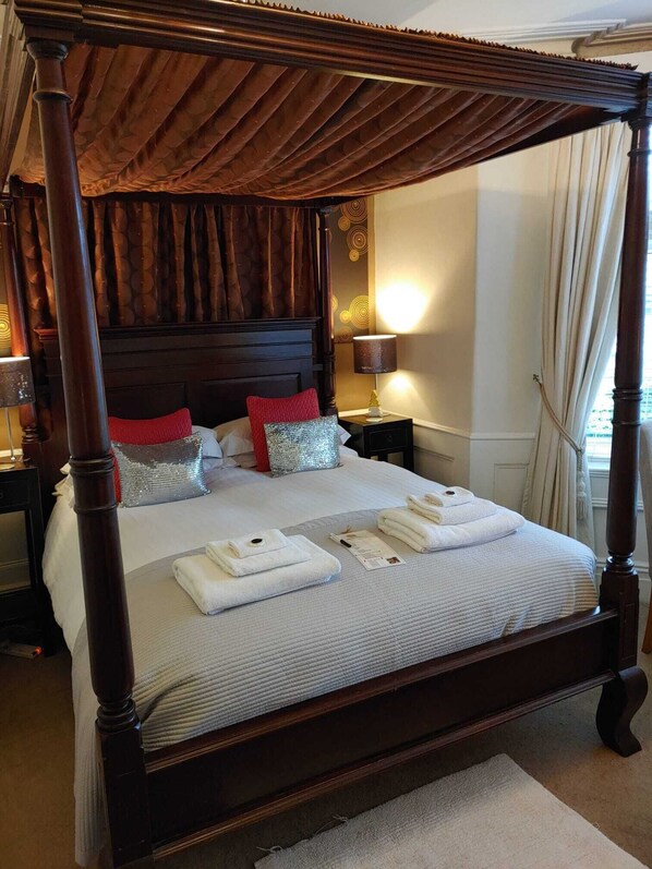 A four-poster bed with a dark wooden frame, white bedding, and red and silver pillows. There are towels on the bed and a lamp on the nightstand. - The Bath House Boutique B&B (Bath)