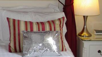 A bed with white bedding, red and beige striped pillows, and a shiny silver pillow. There is a lamp on a bedside table next to the bed.