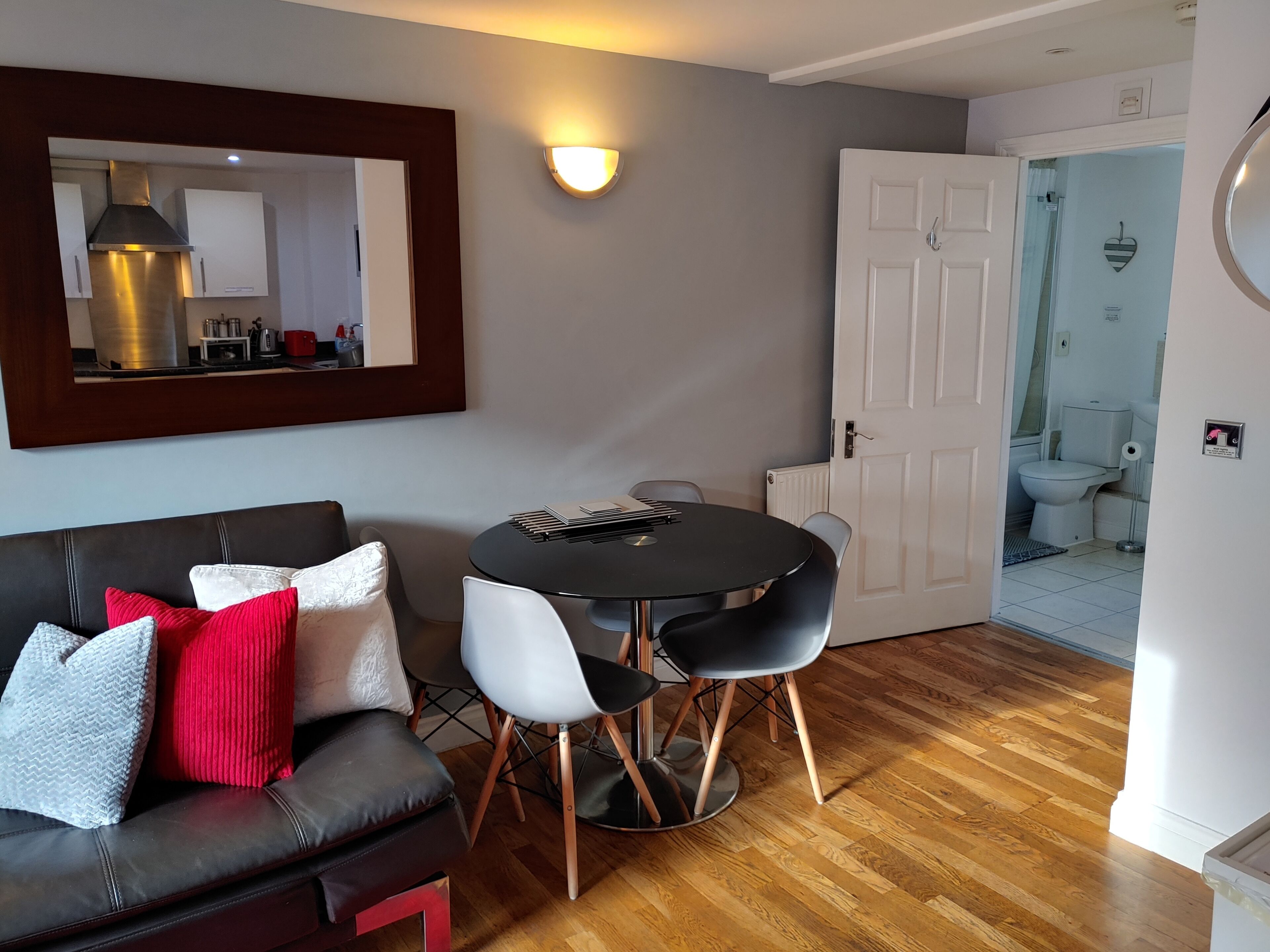 A modern living room with a black round table, white and black chairs, a leather sofa with red and grey cushions, a large mirror, and a white door leading to a bathroom.