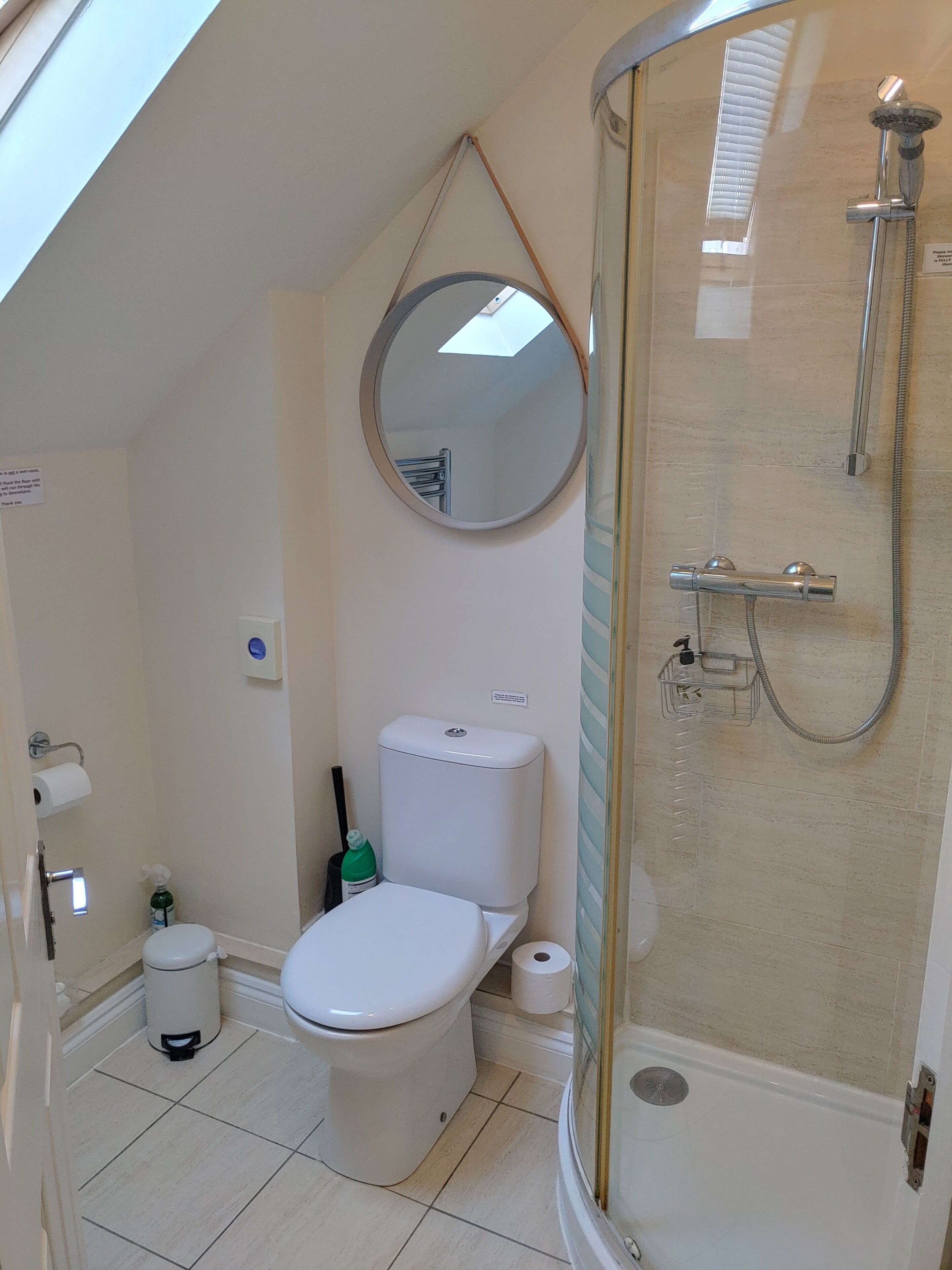 Et badeværelse med toilet, bidet, brusebad med glasdør og et rundt spejl på væggen.