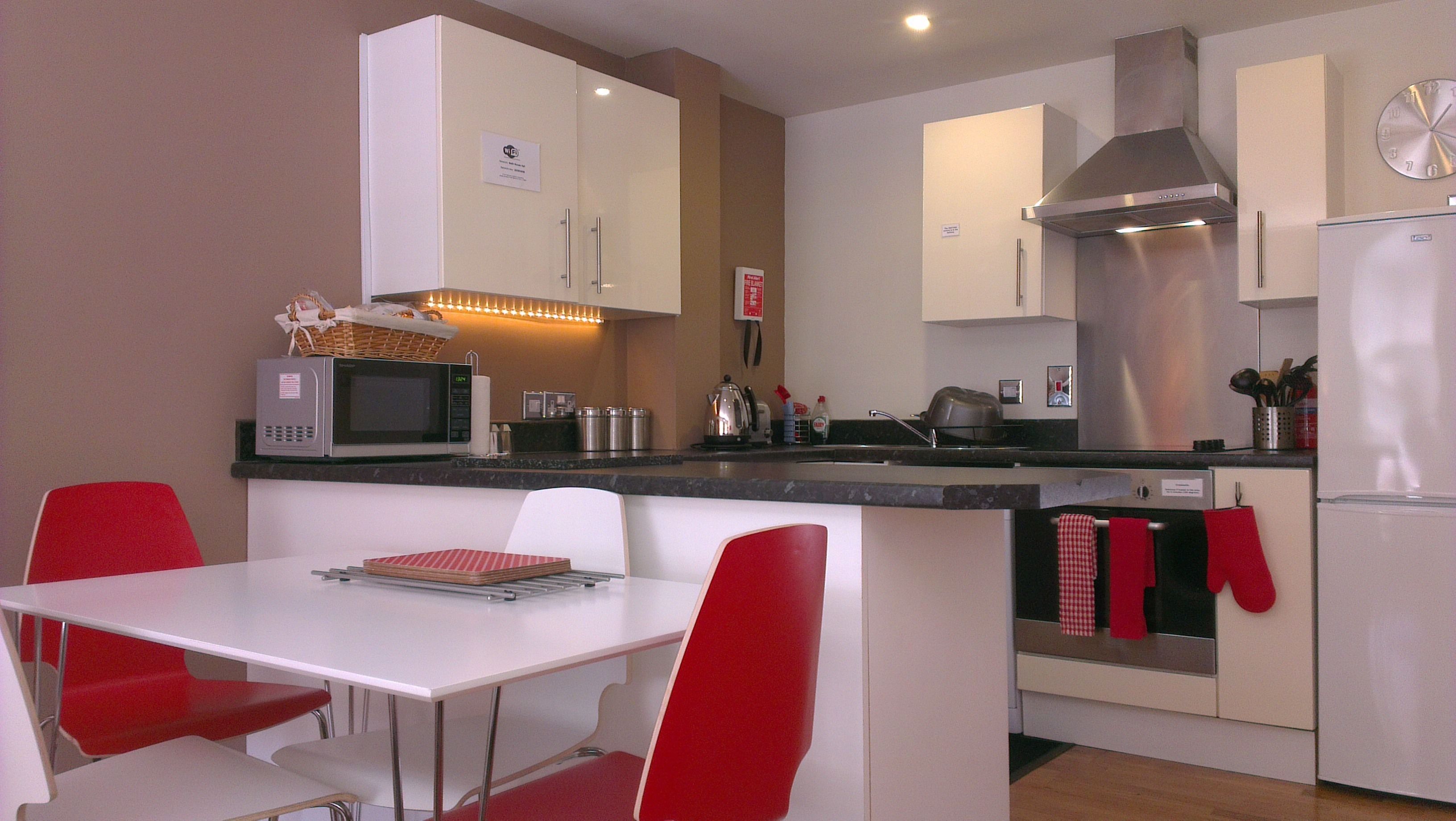 Een compacte keuken met witte kastjes, een magnetron, waterkoker en broodrooster. Er is een eettafel met rode stoelen en een klein keukeneiland met een rood-wit geruit tafelkleed.