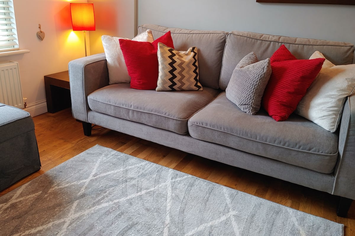 Un sofá gris con varios cojines rojo y beige, una alfombra gris y una lámpara fijada a la pared.