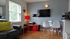 Uma sala de estar moderna com um sofá de couro preto, uma luminária vermelha, uma televisão de tela plana e um espaço de jantar com cadeiras brancas.