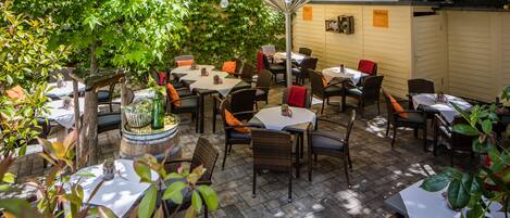 Un patio al aire libre con varias mesas y sillas, rodeado de follaje y cubierto por una sombrilla grande.