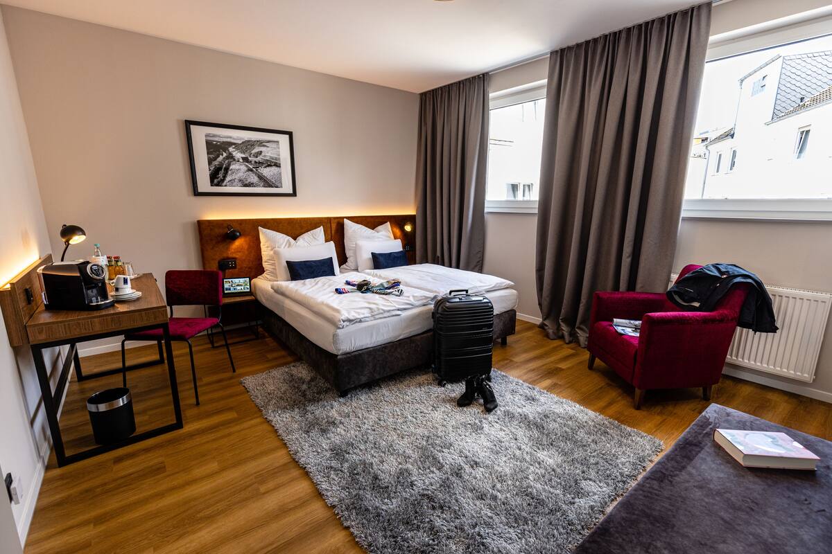 Een hotelkamer met een bed, een bureau, een stoel, een koffer en een raam met gordijnen.