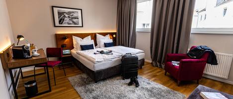 Une chambre d’hôtel avec un lit, un bureau, une chaise, une valise et une fenêtre avec des rideaux.