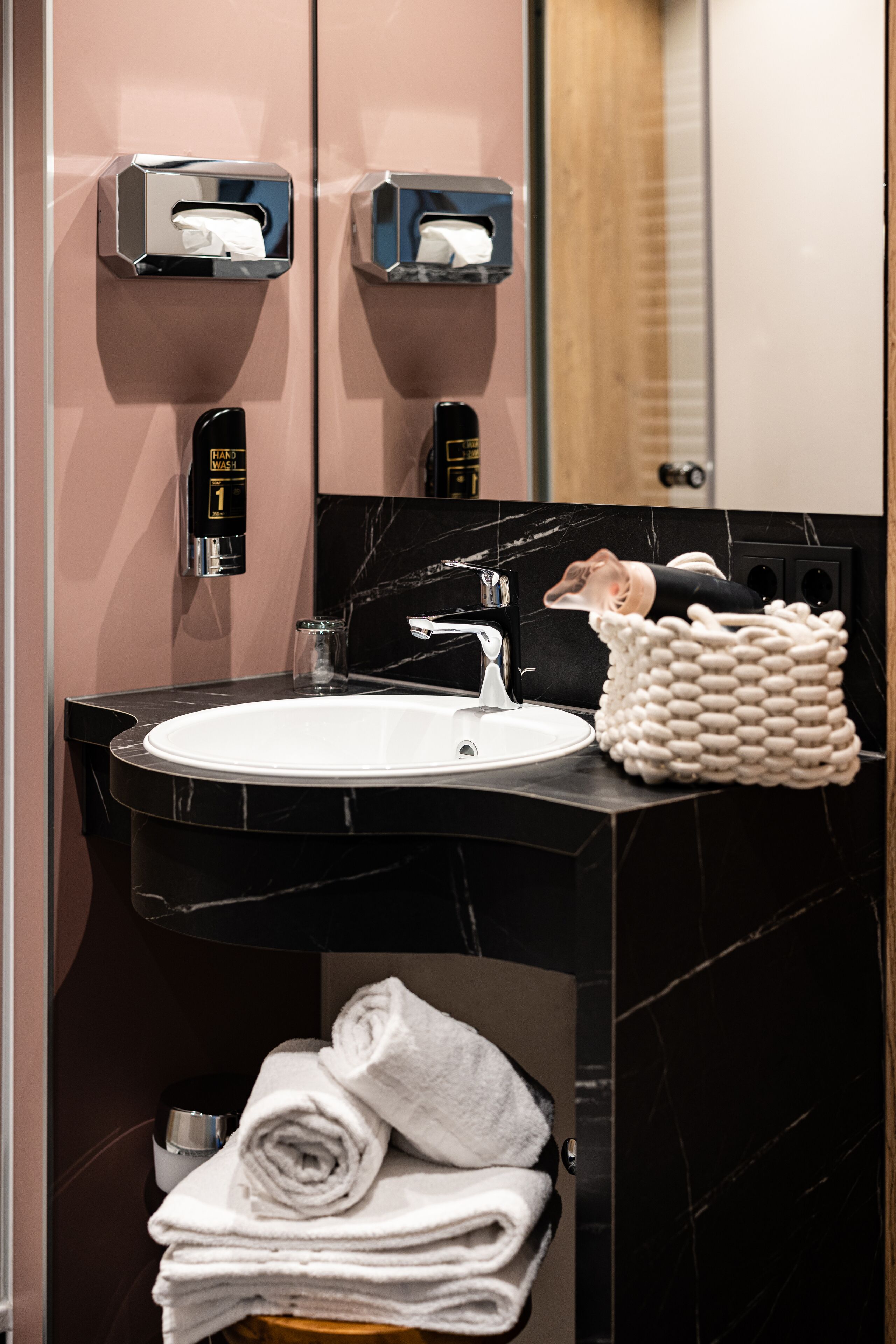Un baño con lavamanos de mármol negro, dos dispensadores de toallas de papel, un secador de manos y un perchero con toallas dobladas.