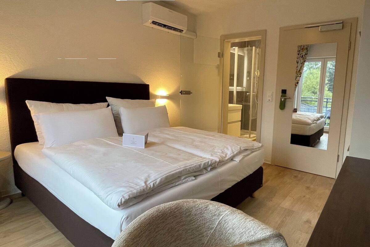 Een net opgemaakt bed met witte lakens, een nachtkastje en een stoel.