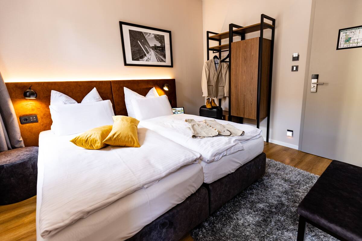 Een hotelkamer met een net opgemaakt bed, een nachtkastje met een lamp, een kapstok en een lijst met een afbeelding aan de muur.