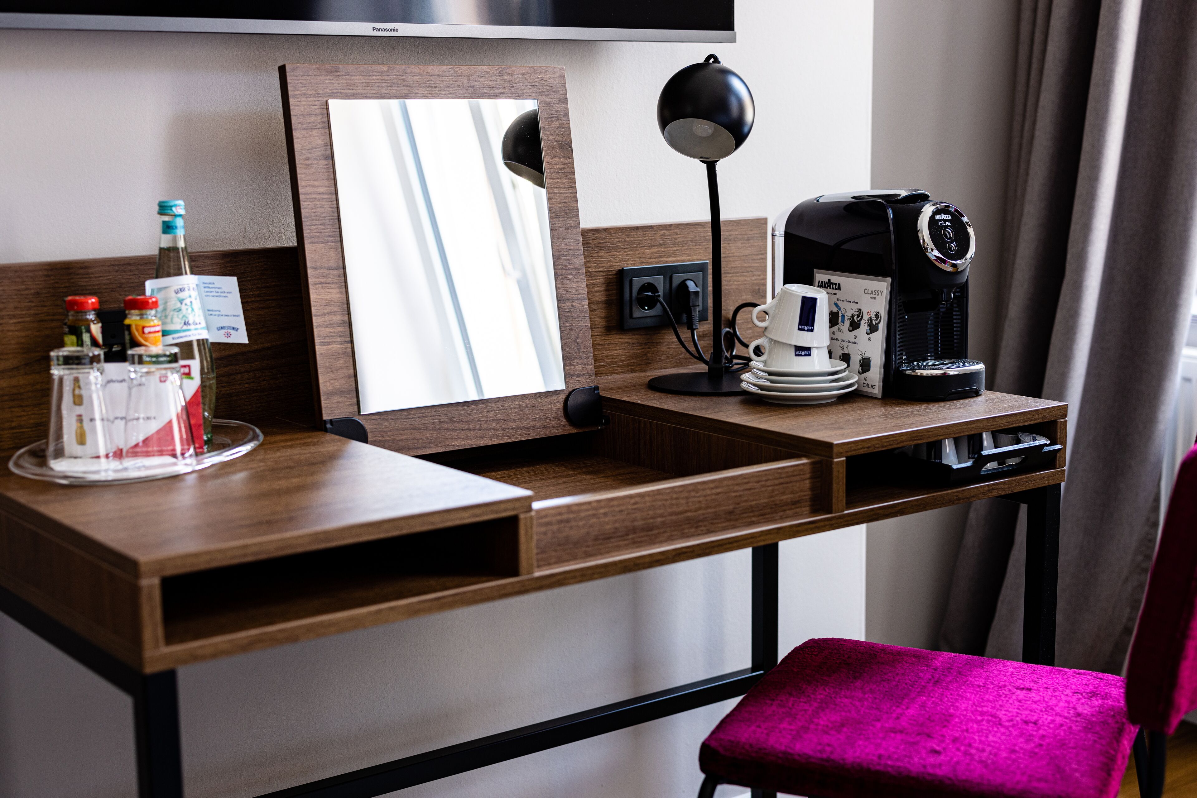 A hotel room with a wooden desk, a coffee maker, a mirror, and a pink chair.