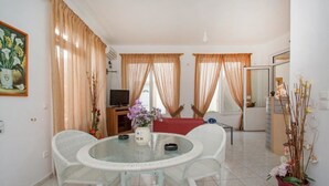 A dining area with a round glass table and white wicker chairs, a television, and a red sofa. - Athena Apartments & Studios (Kithira)