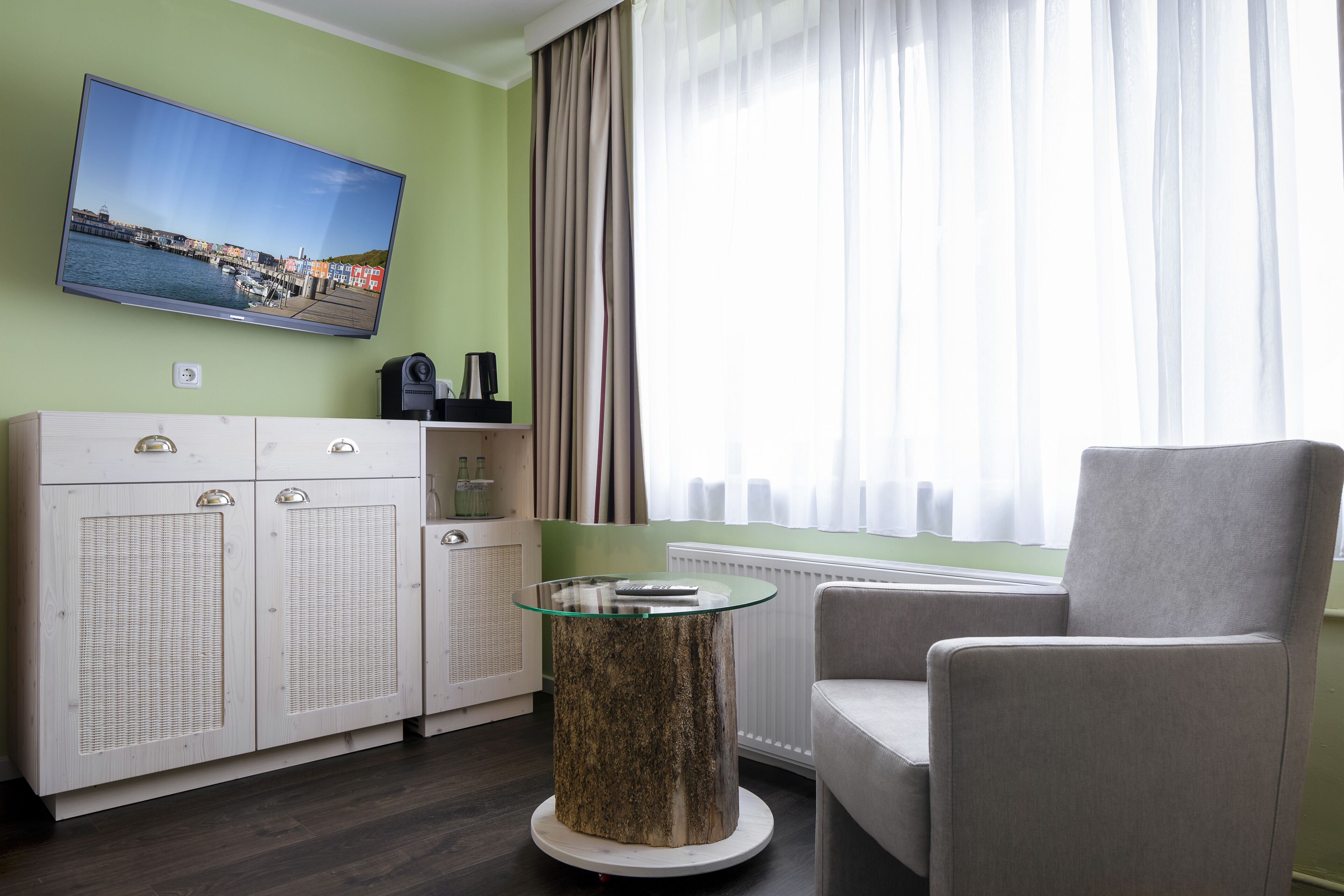 Un salon moderne avec un t éléviseur à écran plat fixé au mur, une table basse ronde au plateau en verre, un fauteuil gris et un meuble blanc à la façade tressée.