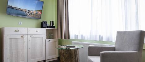 A modern living room with a flat-screen TV mounted on the wall, a round glass-top coffee table, a grey armchair, and a white cabinet with a woven front.