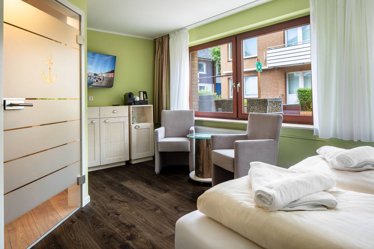 A hotel room with a bed, two armchairs, a small table, a TV, and a view of the outside.