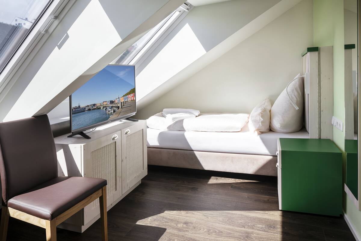 A modern, compact room with a sloped ceiling, a bed, a TV, and a chair.