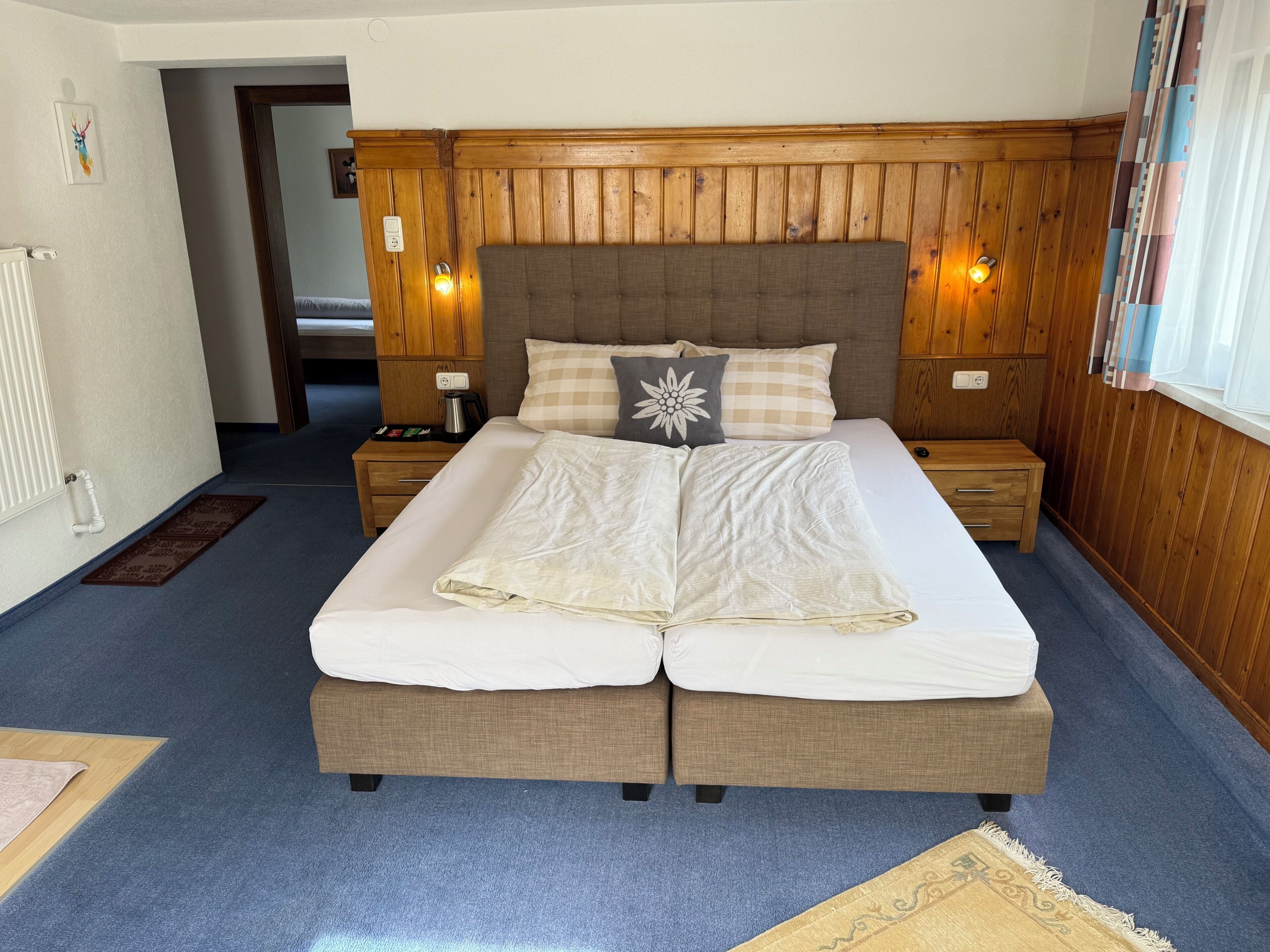 A double bed with a headboard, bedside tables, a nightstand, a radiator, and a door leading to another room.