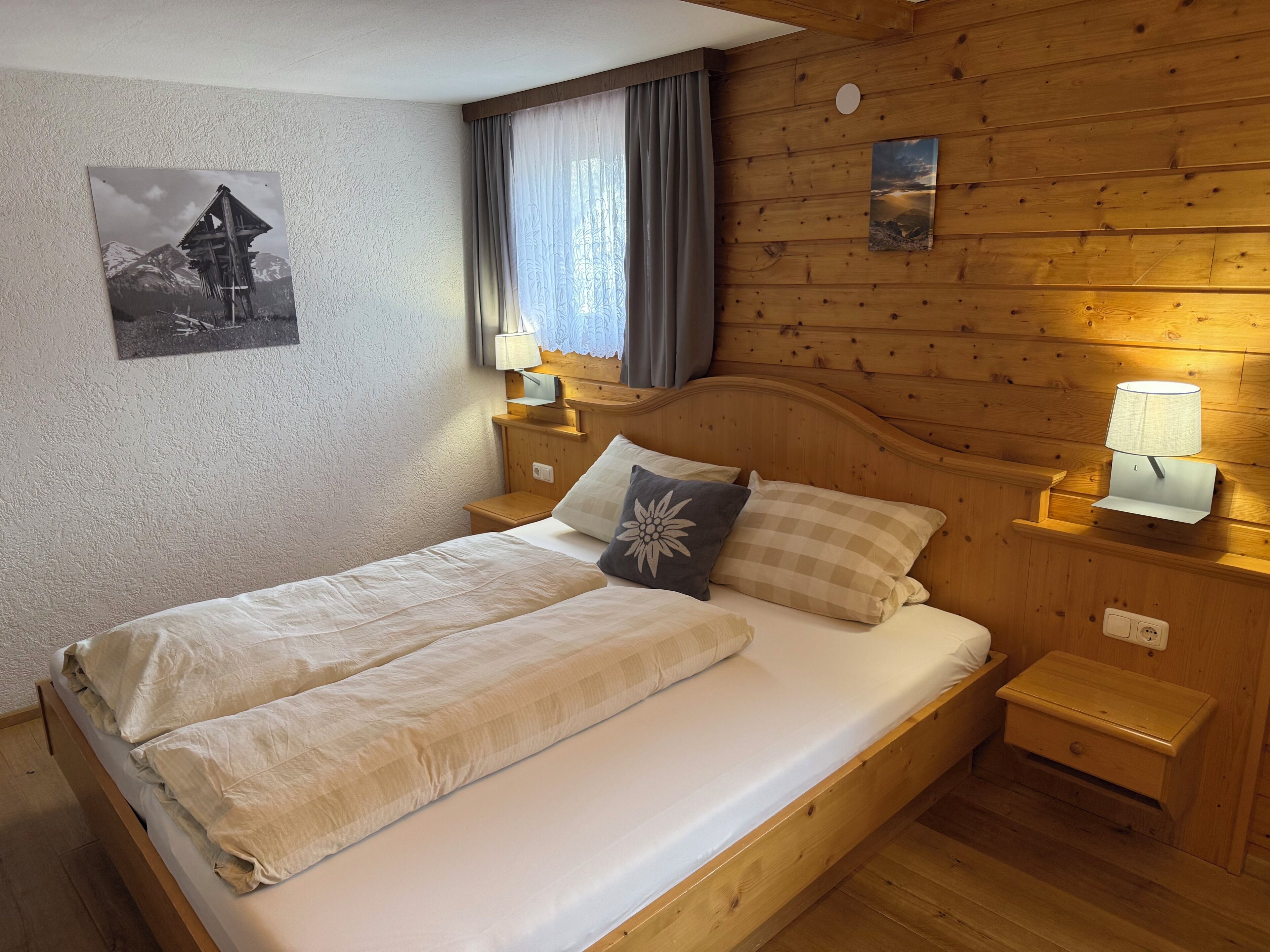 Een houten bed met hoofdeinde, twee nachtkastjes met lampen, een raam met gordijnen en een aan de muur hangend schilderij.