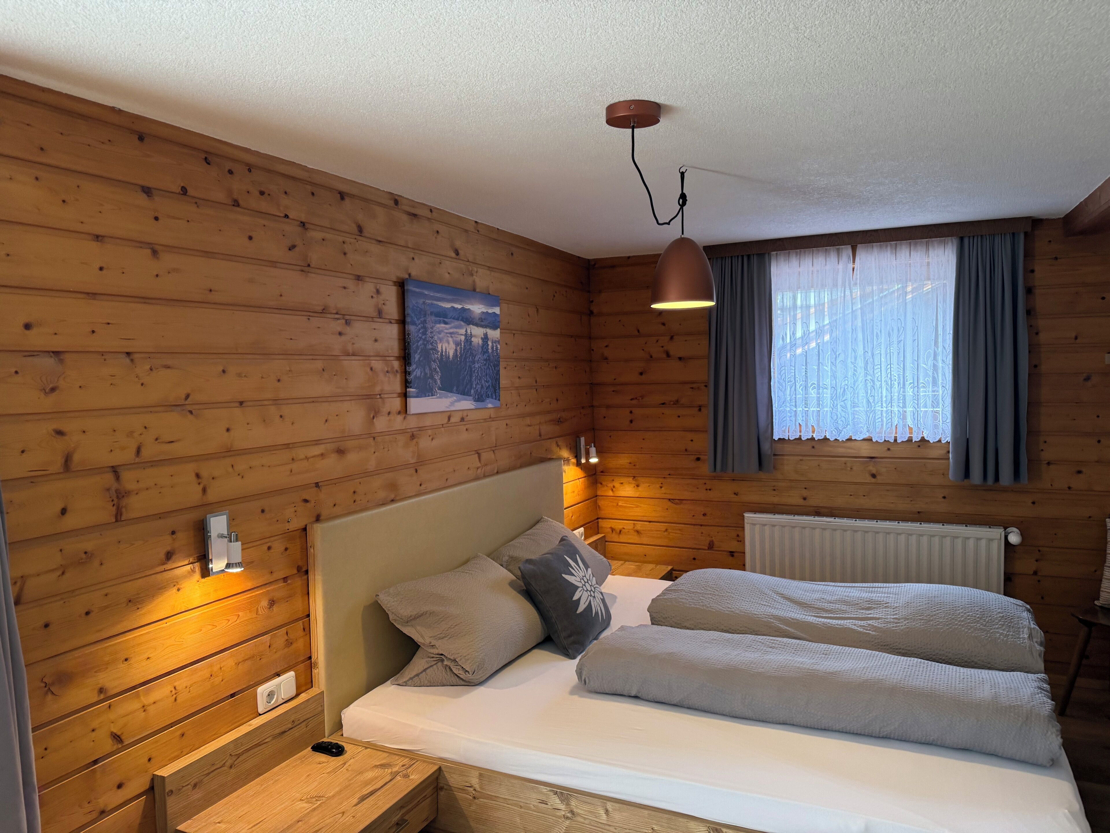 En stuga med träpanel, ett rum med en säng, sänglampor, en tavla och ett fönster med gardiner.