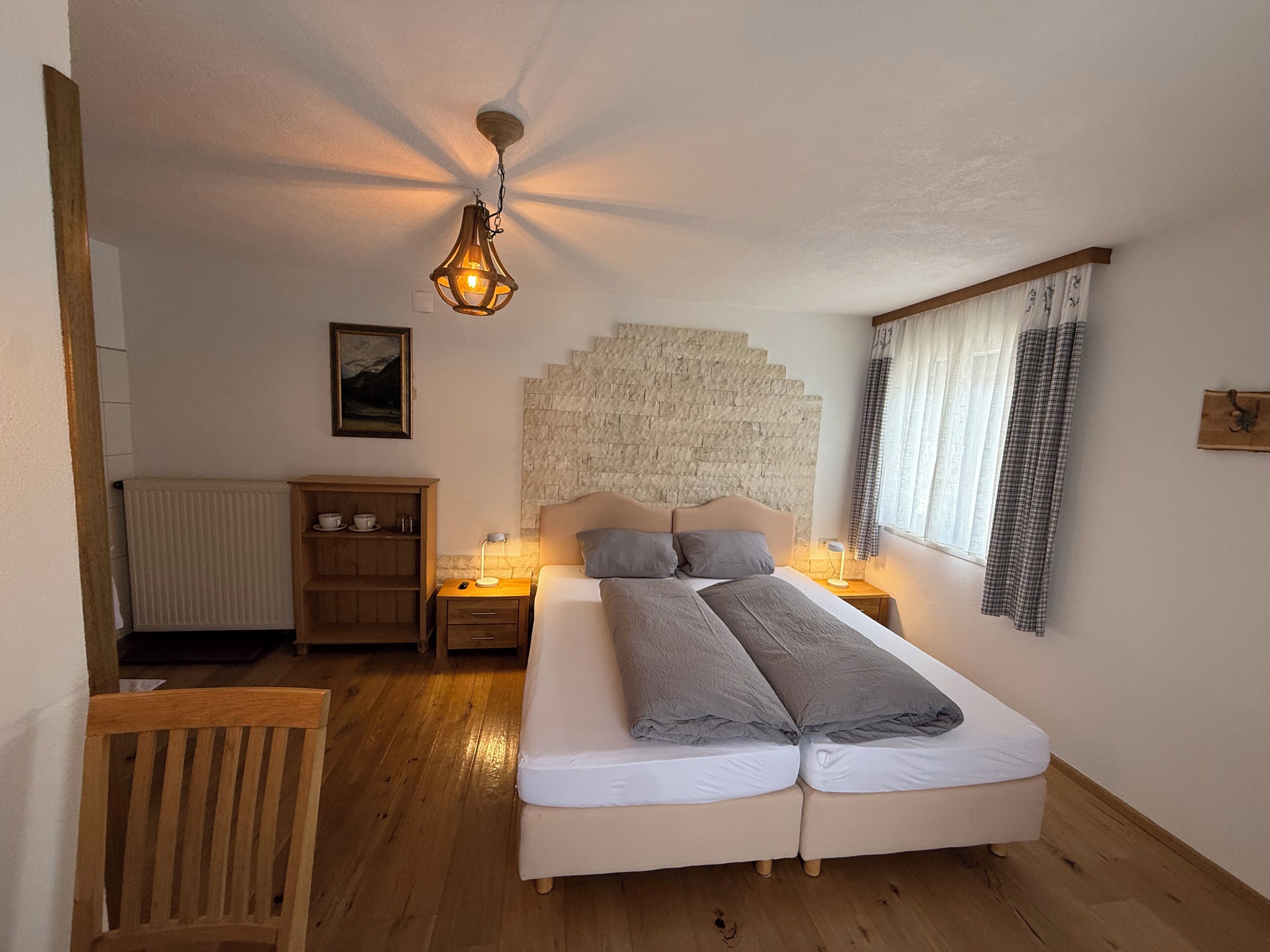 Un dormitorio con cama doble, lámparas de noche, mesita de noche de madera, radiador, silla de madera, pared de piedra y ventana con cortinas.