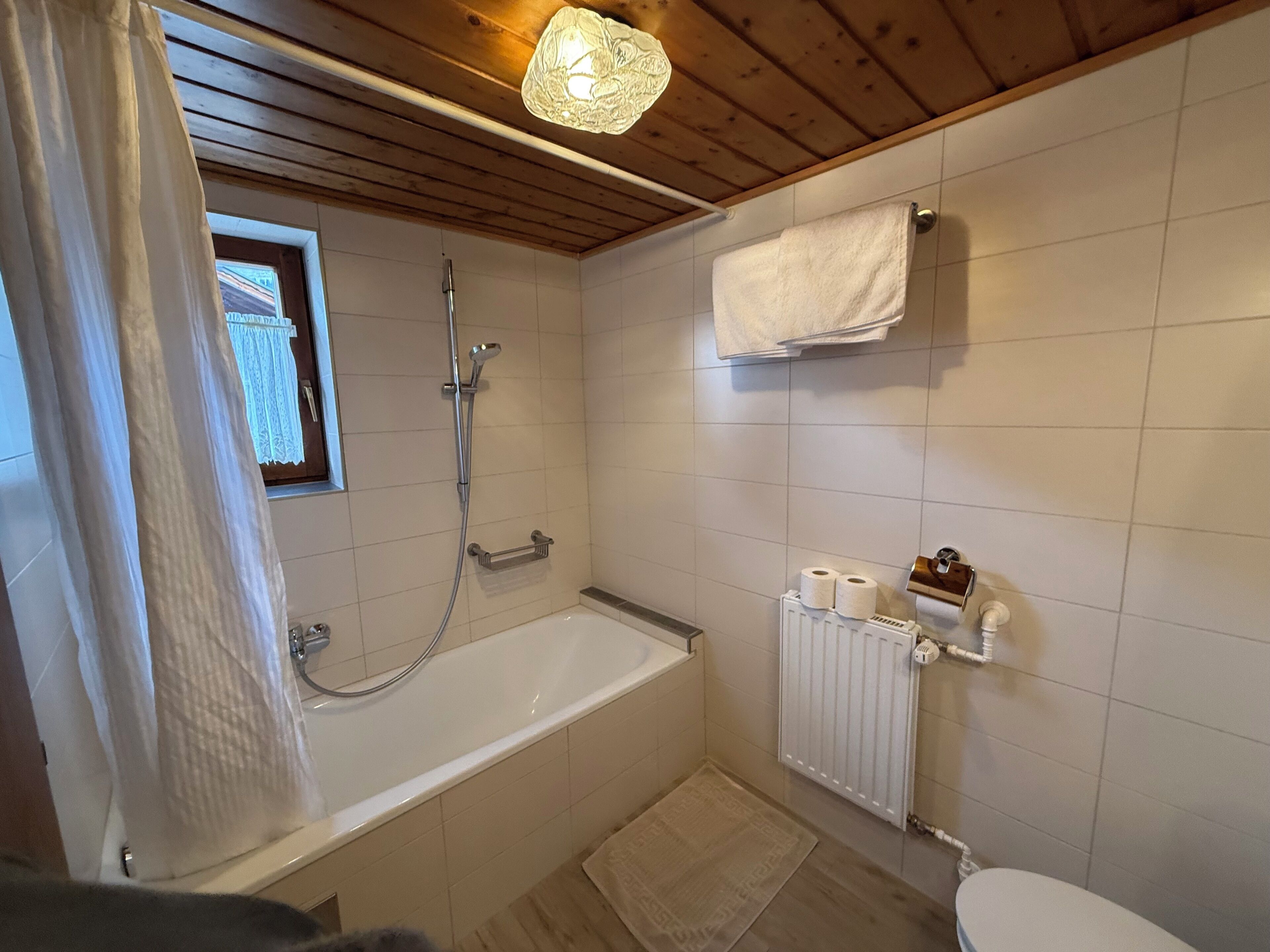 A bathroom with a bathtub, a shower, a toilet, a towel rack, and a window with a curtain.