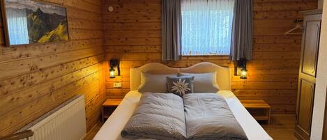 A wooden-paneled room with a double bed, two bedside lamps, a radiator, and a window with curtains.