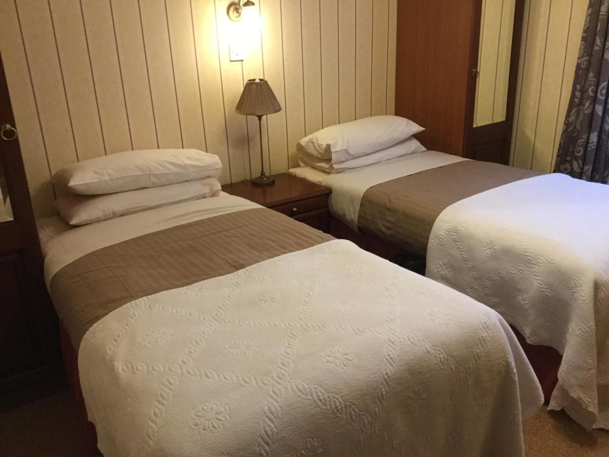 A hotel room with two beds, each with white linens and a brown blanket. There is a bedside lamp on one nightstand and a wooden door.