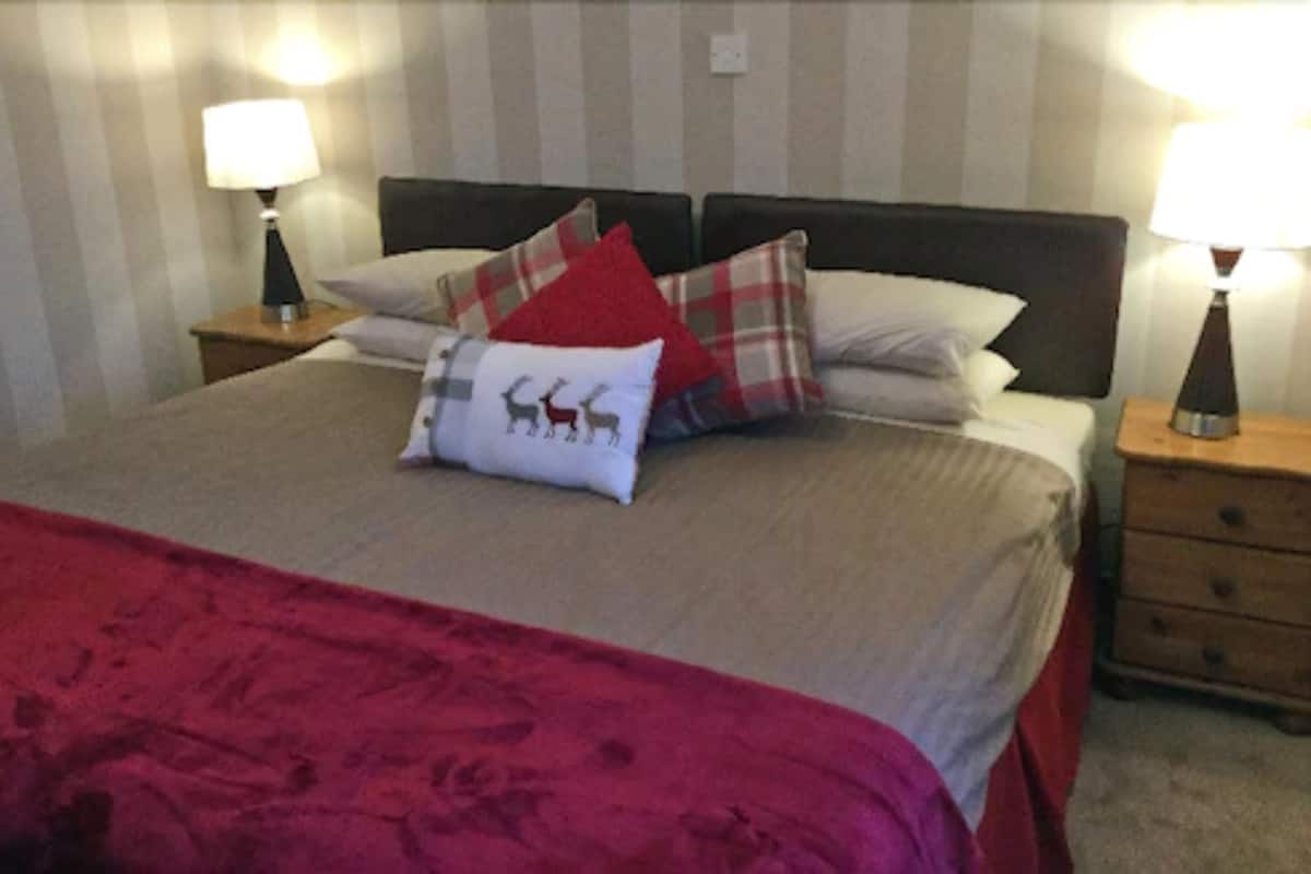 A neatly made bed with a red blanket and multiple pillows, including one with a deer design. There are two bedside tables with lamps, and a wooden nightstand with a drawer.