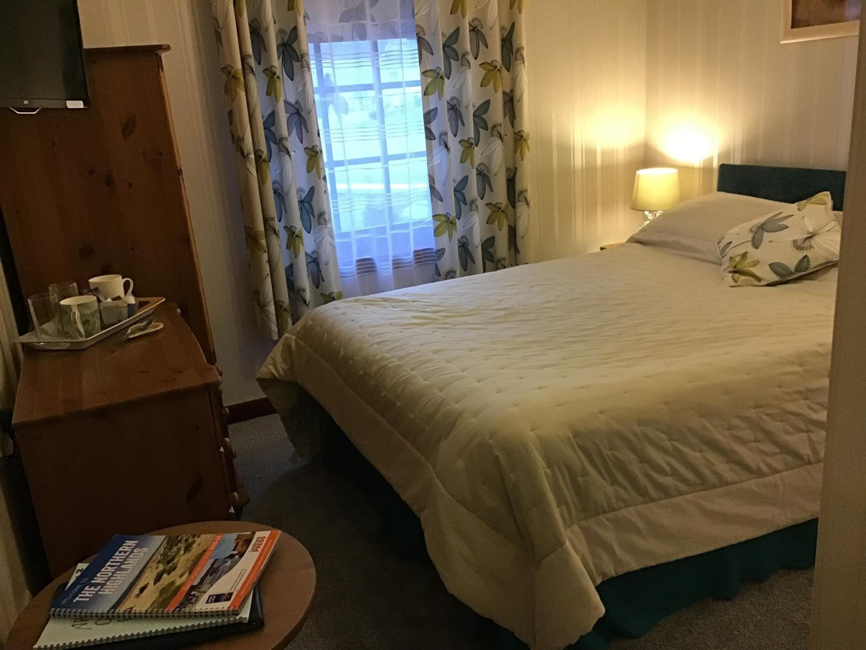 Een hotelkamer met een bed, een nachtkastje met een dienblad ontbijtartikelen, een lamp en een raam met bloemen gordijnen.