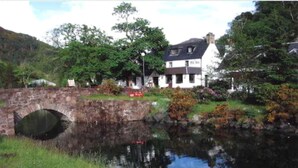 A stone bridge over a calm water body with a house and lush greenery in the background. - The Old Inn (Gairloch)