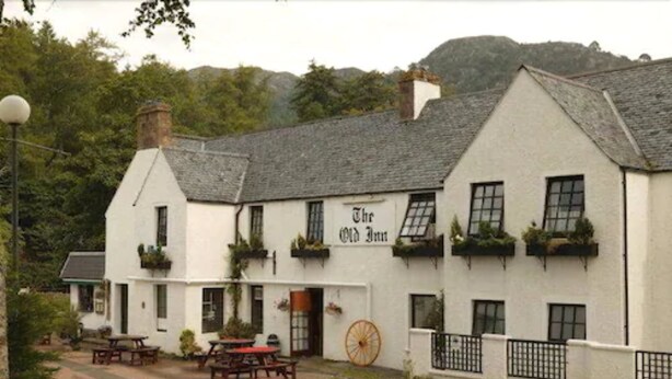 A white building with the sign "The Old Inn" on the front. It has multiple windows, some with plants, and a large wheel in front. - The Old Inn (Gairloch)