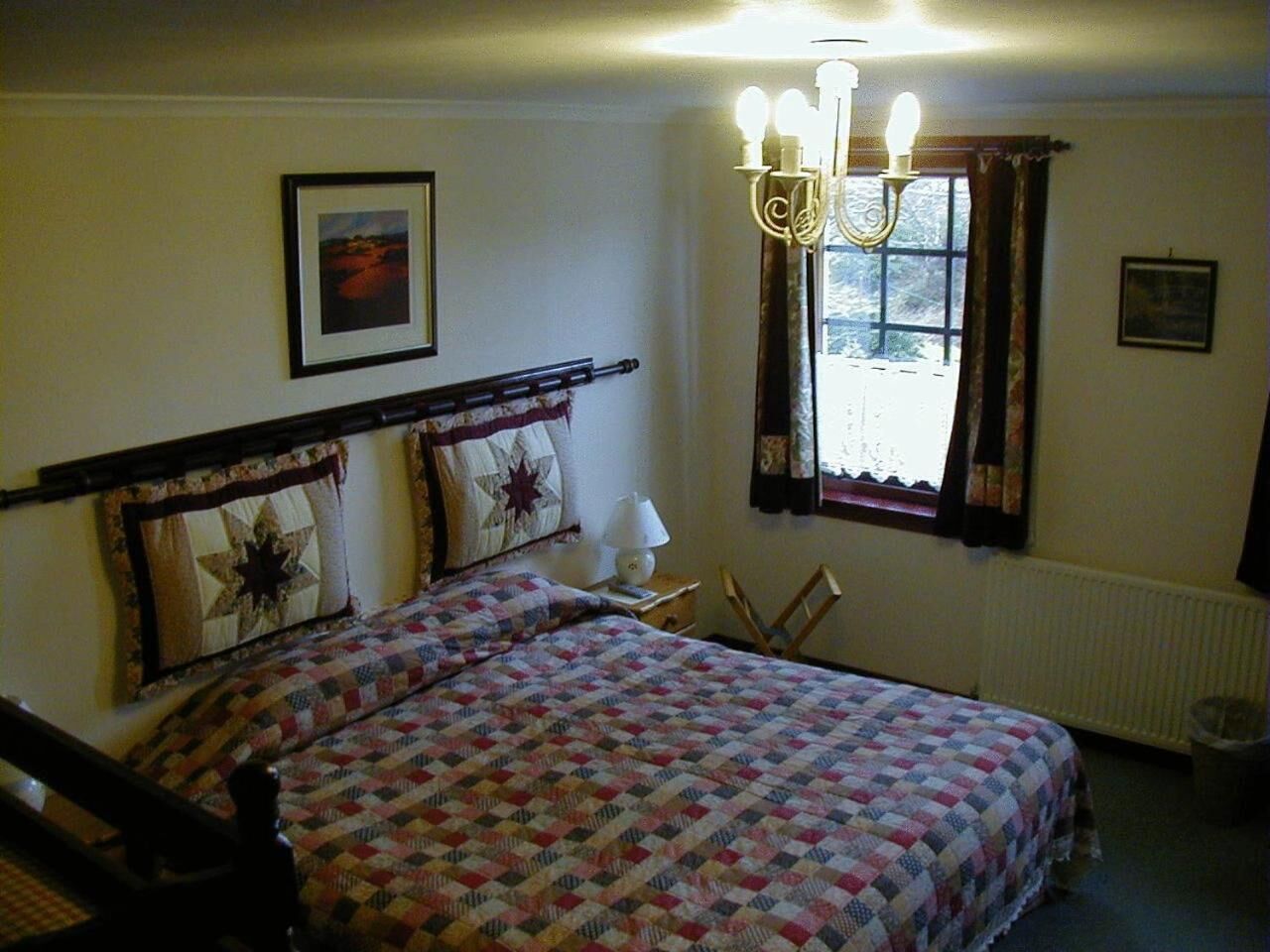 Ein Schlafzimmer mit Bett, Nachttisch, Sessel, Fenster mit Vorhängen und einer Deckenleuchte.