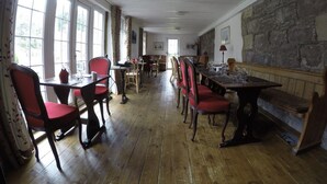 A dining area with wooden tables and chairs, large windows, and a stone wall. - The Old Inn (Gairloch)