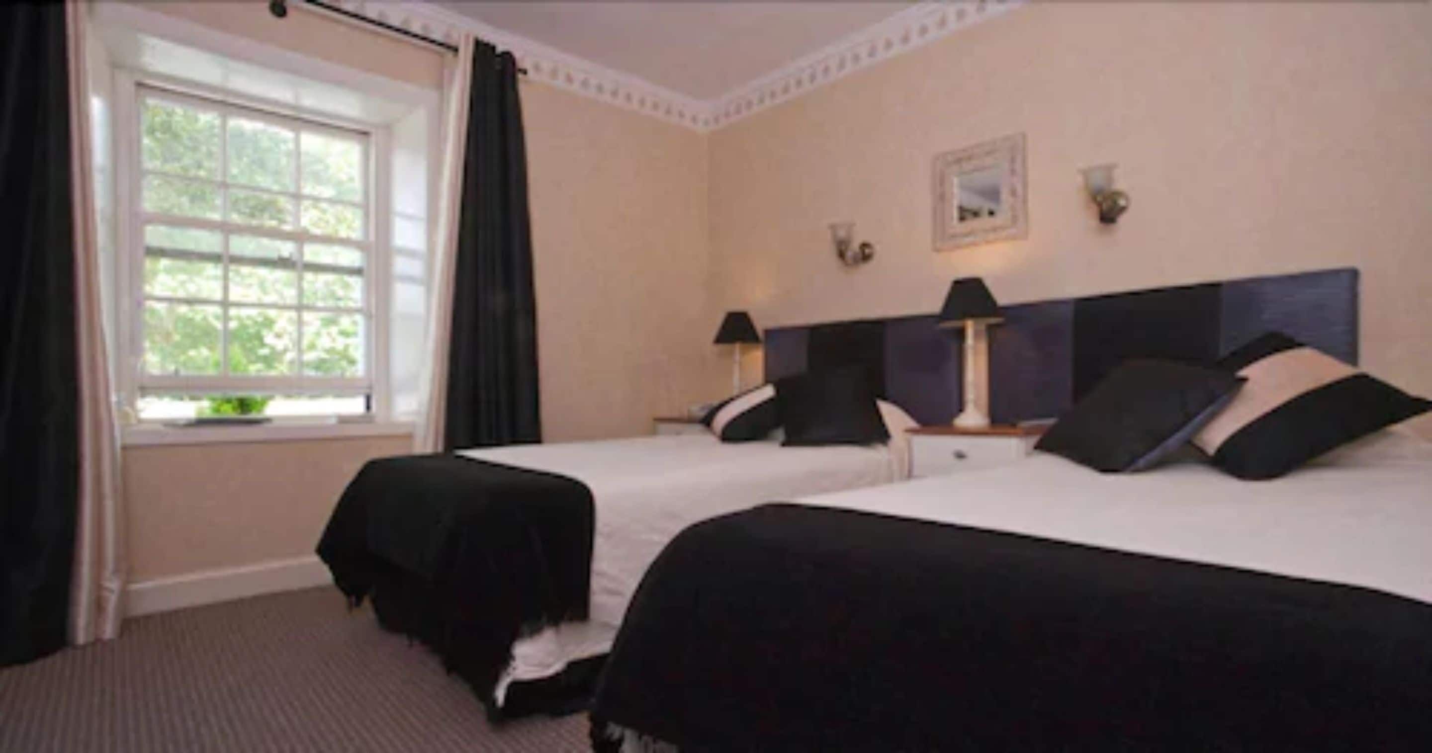 A hotel room with two beds, a window with curtains, a nightstand with a lamp, and a wall-mounted picture.