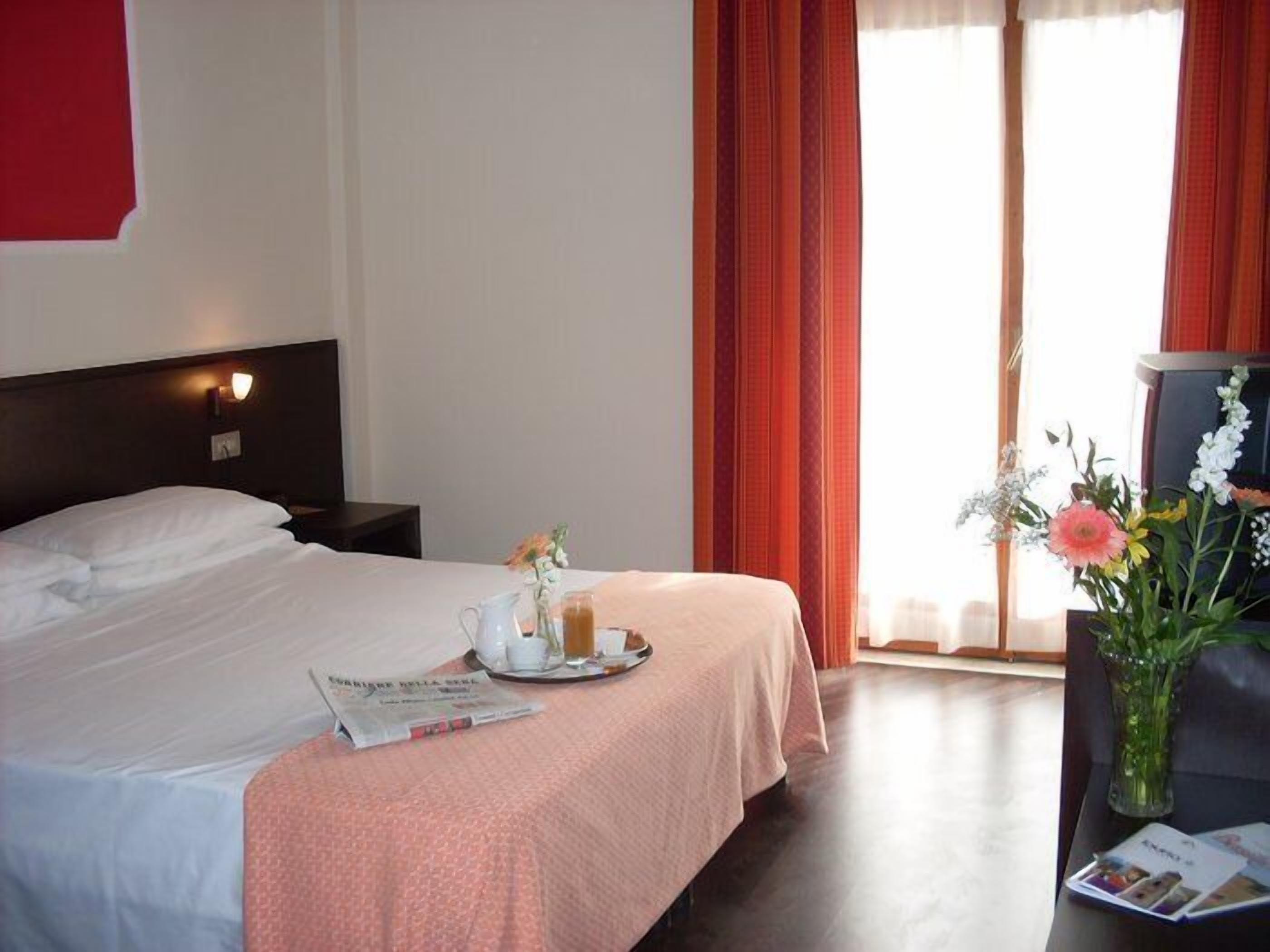 Een hotelkamer met een bed, een nachtkastje, een vaas met bloemen en een raam met gordijnen.