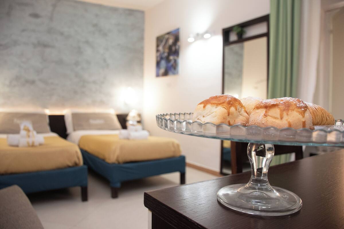 A hotel room with two beds, a glass cake stand with pastries, and a modern decor.