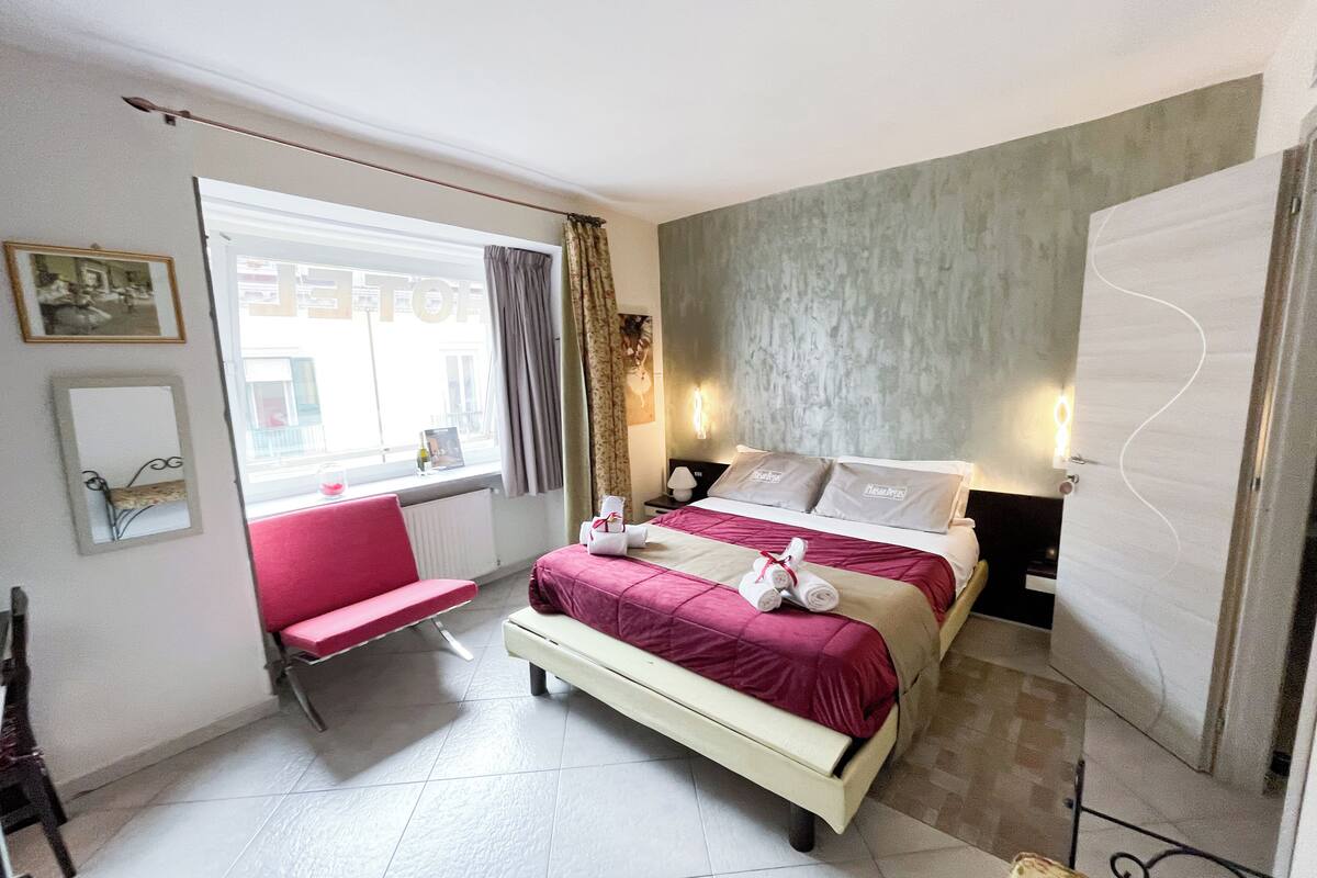 A hotel room with two beds, a red chair, a window with curtains, a radiator, and a wall-mounted light fixture.