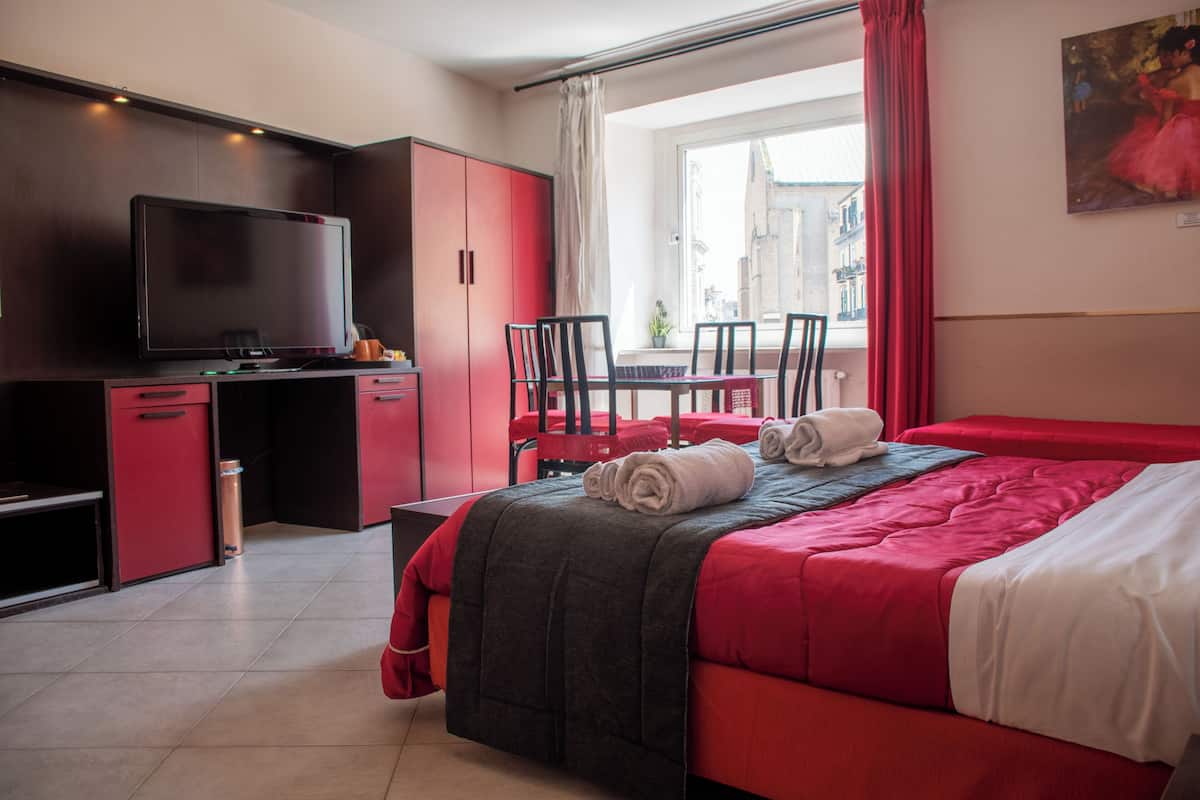 A hotel room with a bed, a desk with a TV, a chair, and a window with curtains.