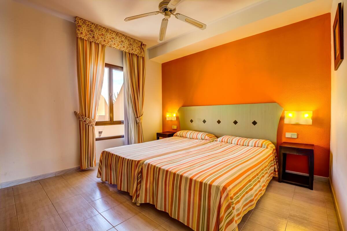 A hotel room with a bed, two bedside tables, a ceiling fan, and a window with curtains.