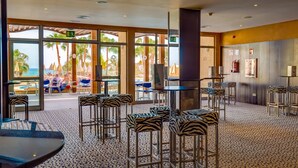 A spacious indoor area with high ceilings, large windows offering views of palm trees and a beach, and zebra-striped bar stools and tables.