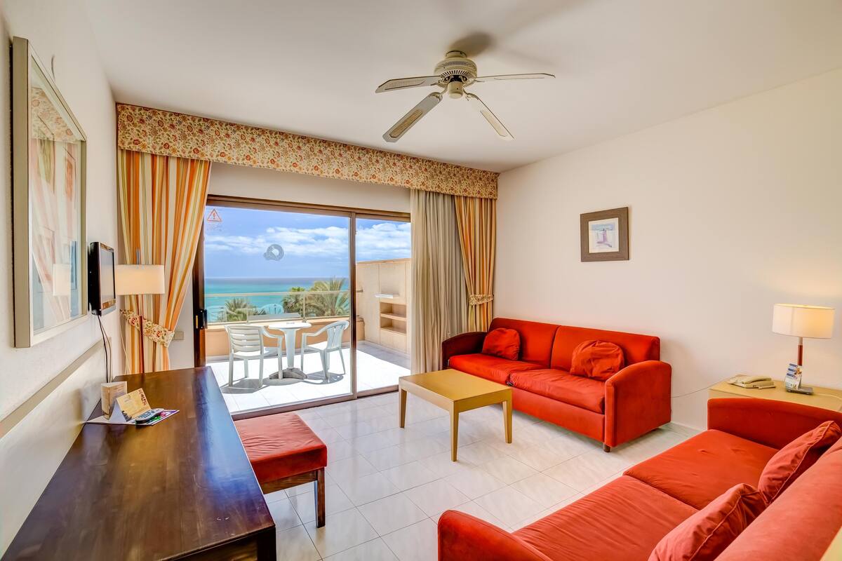 A hotel room with a red sofa, a wooden table, a ceiling fan, and a view of the ocean through a sliding glass door.