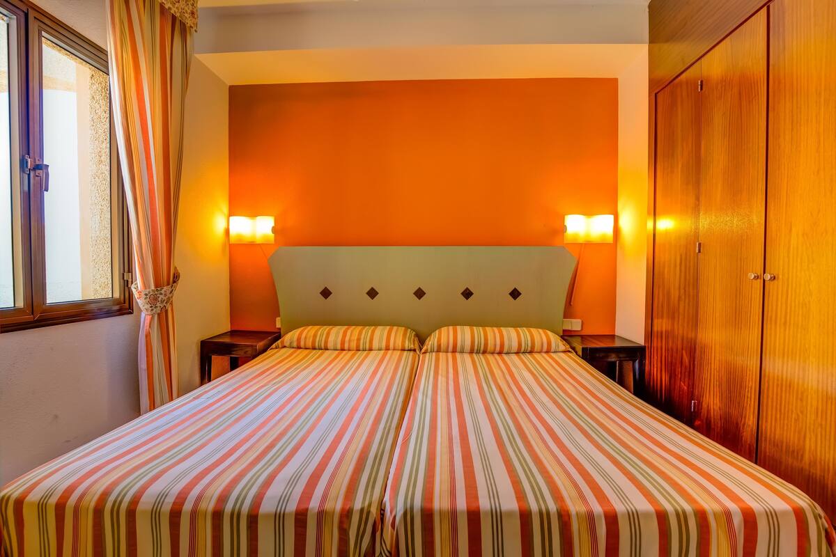 A double bed with striped bedding, two bedside tables, two wall-mounted lamps, and a wooden wardrobe.