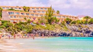 A beachfront hotel with multiple balconies, lush greenery, and a rocky shoreline.