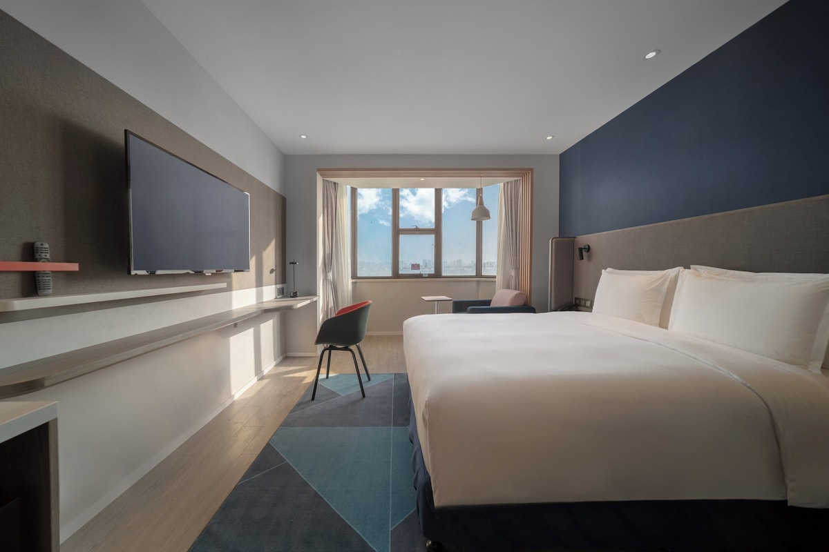 Habitación de hotel con cama, televisión montada en la pared, escritorio y ventana con cortinas.