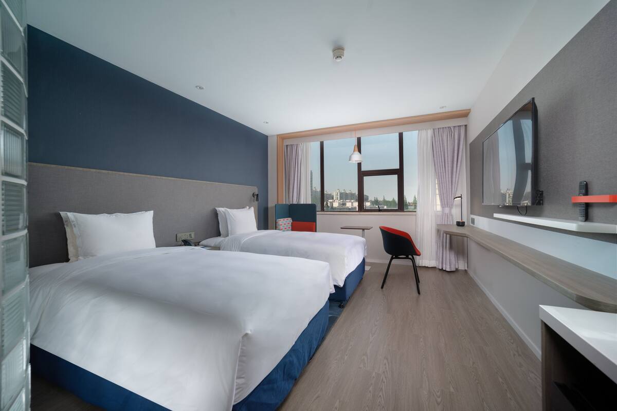 Ett hotellrum med två sängar, ett skrivbord, en stol, en TV och ett stort fönster med utsikt över staden.