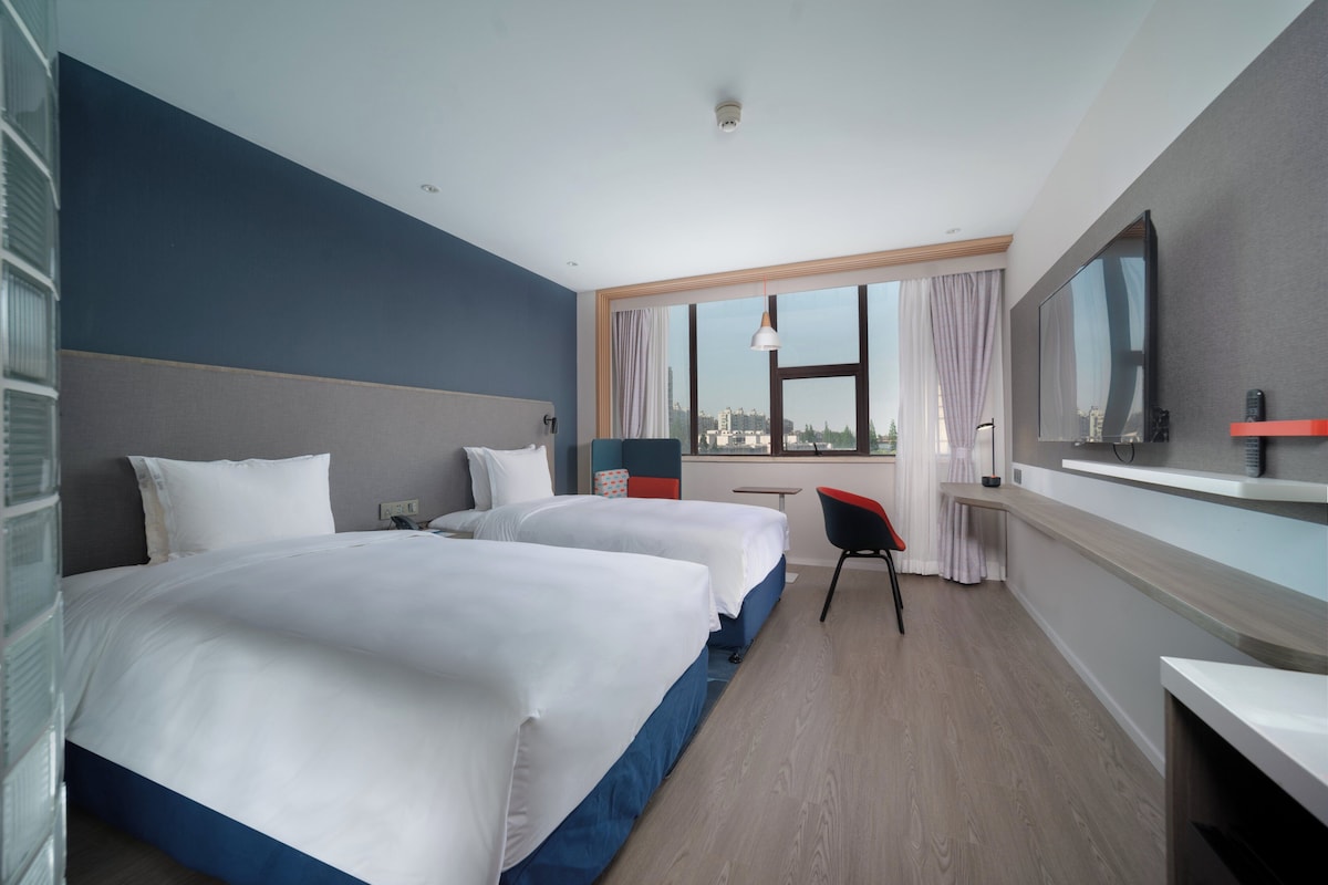 Habitación de hotel con dos camas, un escritorio, una silla, un televisor y un ventanal con vistas a la ciudad.