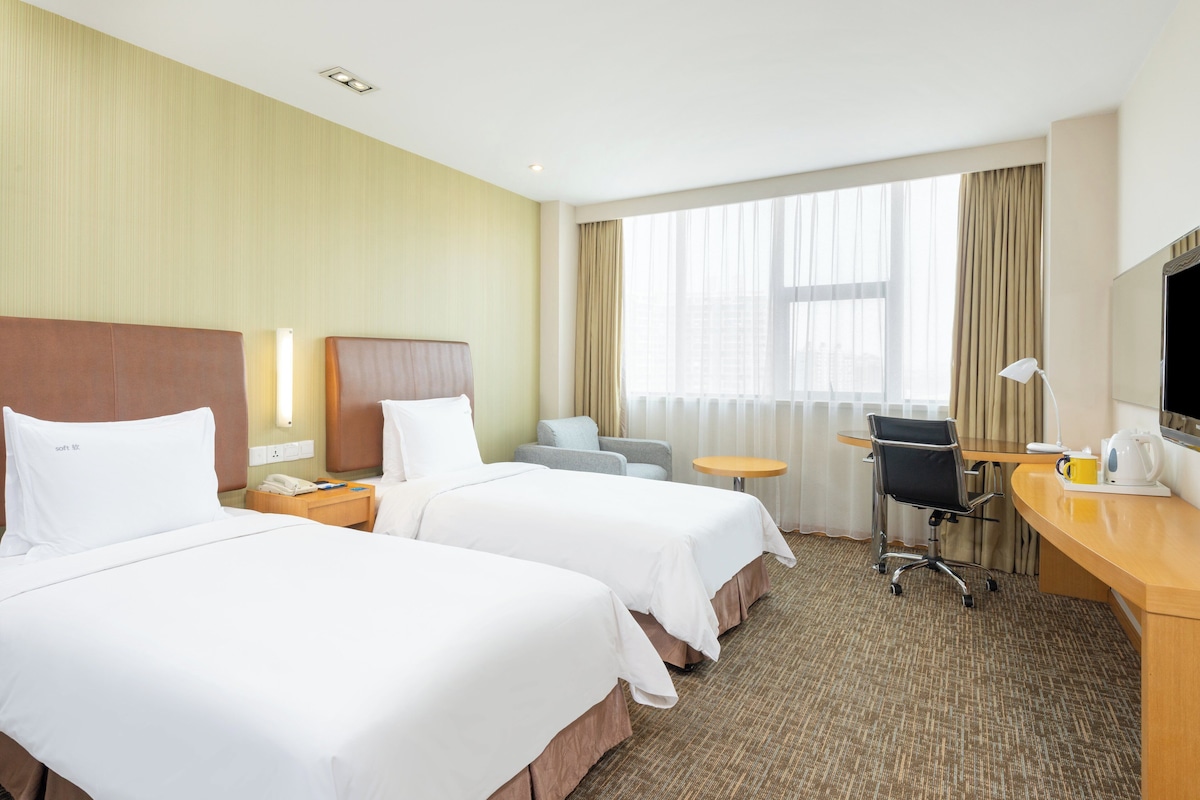 Habitación de hotel con dos camas, un escritorio con silla, una mesita redonda y un gran ventanal con cortinas.