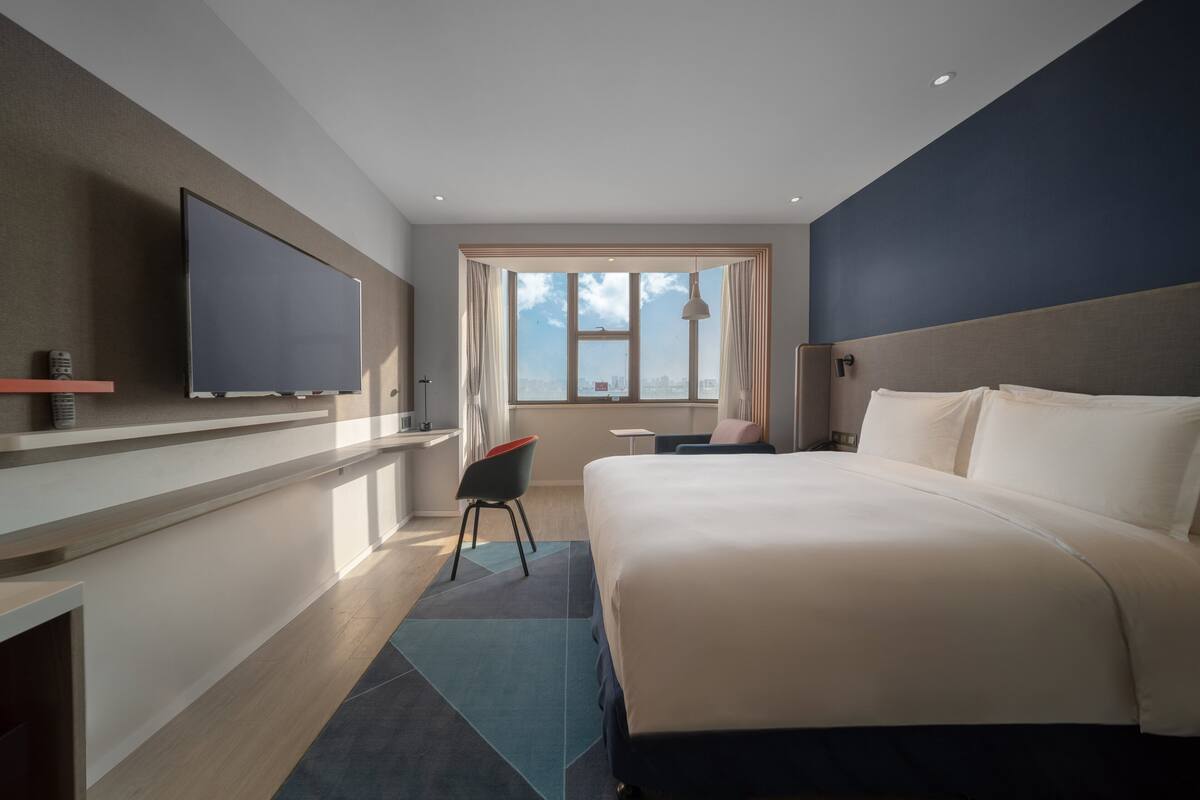 Ett modernt hotellrum med en stor säng, en platt-TV som är monterad på väggen, ett skrivbord med en stol och ett fönster med utsikt över staden.