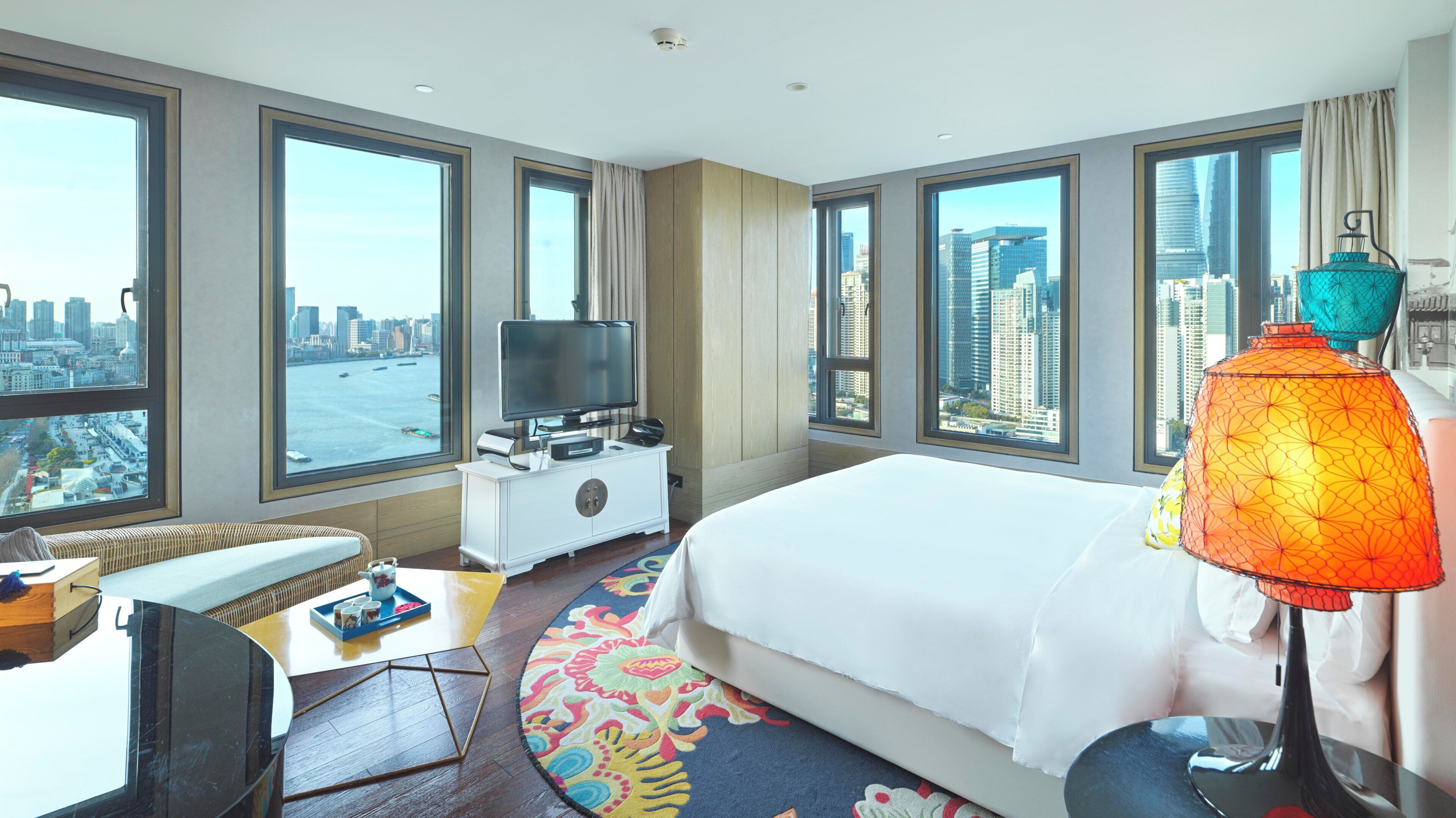 A hotel room with a large bed, a TV, a desk, and a view of the city and water.