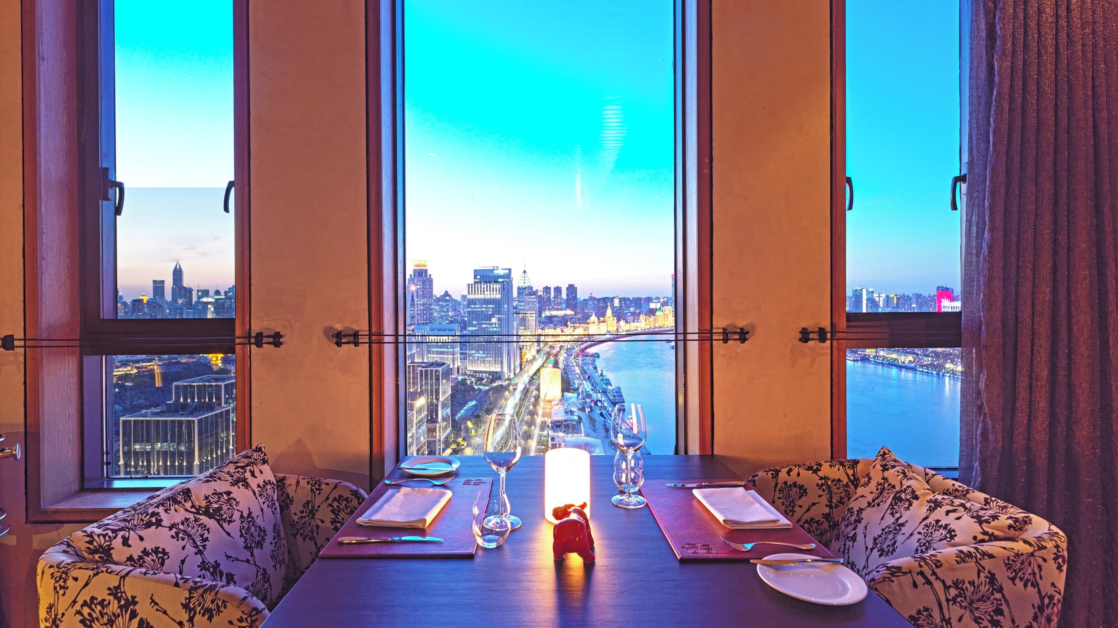 Un comedor con una mesa dispuesta para dos personas, ventanas amplias con vistas a la ciudad y una vela decorativa sobre la mesa.